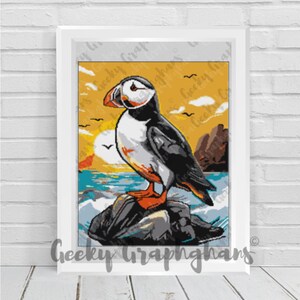May include: A framed print featuring a colorful illustration of a puffin bird perched on a rock. The artwork showcases the puffin with its distinctive orange beak, set against a backdrop of a sunset over the ocean. The text "Geeky Graphghans" is visible.