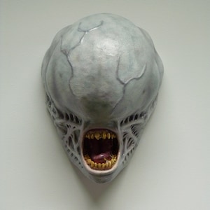 Könnte beinhalten: Eine graue Alien-Maske mit weit offenem Mund und scharfen Zähnen. Die Maske ist aus einem strukturierten Material gefertigt und hat ein realistisches Aussehen.