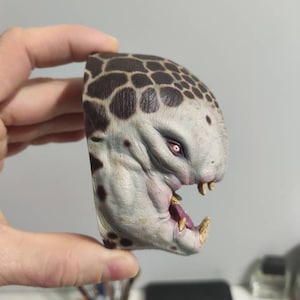 May include: Close-up side view of a painted miniature creature with a spotted, grey and brown skin tone. The creature has a large, open mouth with sharp teeth and a single red eye.