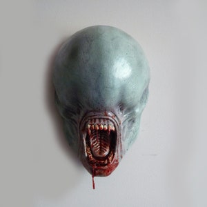 Könnte beinhalten: Eine graue Alien-Kopfskulptur mit einem großen, offenen Mund und tropfendem Blut. Die Skulptur ist aus glattem, bemaltem Material gefertigt und hat ein realistisches Aussehen.