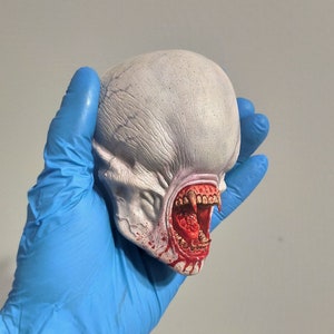 Könnte beinhalten: Ein weißer, detaillierter, geformter Kopf mit einem großen, offenen Mund, der scharfe Zähne und rotes Blut zeigt, das aus dem Mund tropft.