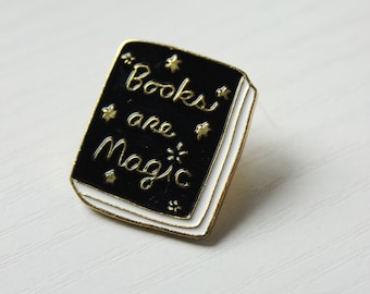 May include: Black enamel pin shaped like a book with gold trim and the text "Books are Magic" in gold lettering.