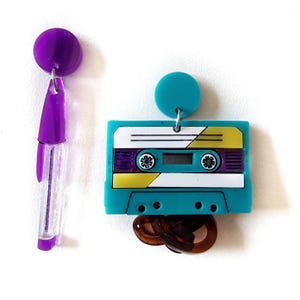 May include: A pair of dangle earrings. One earring features a purple pen-shaped charm and a purple circle. The other earring features a turquoise cassette tape charm with a yellow and purple design, and a brown chain charm.