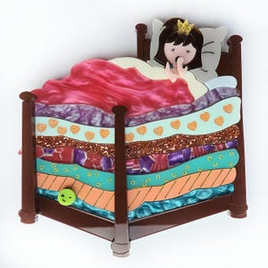 以下が含まれることがあります： アクリルを重ねて作られた、ベッドにいるお姫様を描いた気まぐれなブローチ。ベッドは茶色で、ピンク、紫、ティール、オレンジ色のカラフルな寝具の層があります。お姫様は金の王冠をかぶり、あくびをしています。小さな緑色の笑顔が側面にあります。