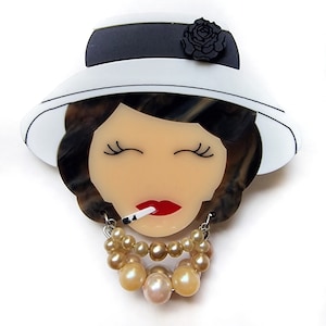 Può includere: Un cappello bianco con una fascia nera e una rosa nera sul lato. Il cappello è indossato da una donna con i capelli castani e gli occhi chiusi. Indossa una collana di perle e ha una sigaretta in bocca.