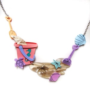 May include: A colorful necklace featuring a pink bucket, a purple shovel, a blue seashell, a brown seashell, a green seahorse, a purple turtle, and two turquoise starfish.