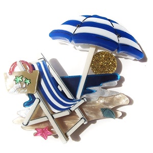 May include: A blue and white striped beach chair with a matching blue and white striped umbrella. The chair has a tan and white striped cushion and a tan bag with palm trees on it. The umbrella is over a gold glitter sun and a blue and white beach scene.