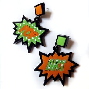 Könnte beinhalten: Ein Paar schwarze, orange und grüne Statement-Ohrringe mit Pop-Art-Design. Die Ohrringe haben eine schwarze Sternform mit den Worten "POP" und "ART" in orangefarbenen Buchstaben mit weißen Punkten auf grünem Hintergrund.