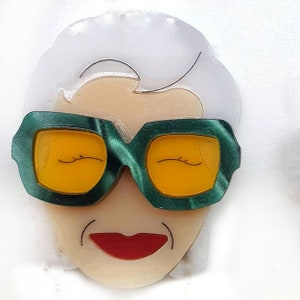 Può includere: Un broche bianco e verde con un viso di cartone animato che indossa occhiali da sole verdi e una bocca rossa.
