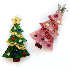 Può includere: Due spille a forma di albero di Natale, una verde con glitter dorati e una rosa con glitter rossi. Entrambe hanno una stella in cima e sono decorate con palline.