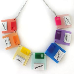 Puede incluir: Un collar colorido con colgantes de disquetes de plástico en varios colores, incluyendo rosa, naranja, amarillo, verde, azul y morado. Cada disco tiene una pequeña etiqueta con texto como "RAINBOW.EXE" y "DISK 1". El collar tiene una cadena plateada.