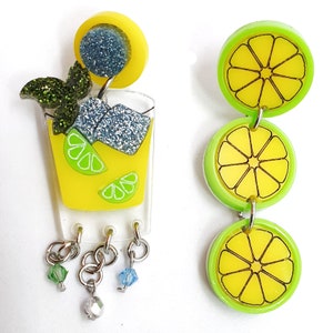 May include: A pair of novelty earrings. One is a yellow cocktail with glitter ice and mint, the other has three yellow lime slices with green rims. Both earrings have dangling charms. The earrings are made of acrylic.