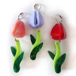 May include: Three pairs of flower-shaped earrings. Each earring has a colourful tulip-like top in red, pink, or purple, connected to a green stem with green leaves. The earrings have silver-coloured ear wires.