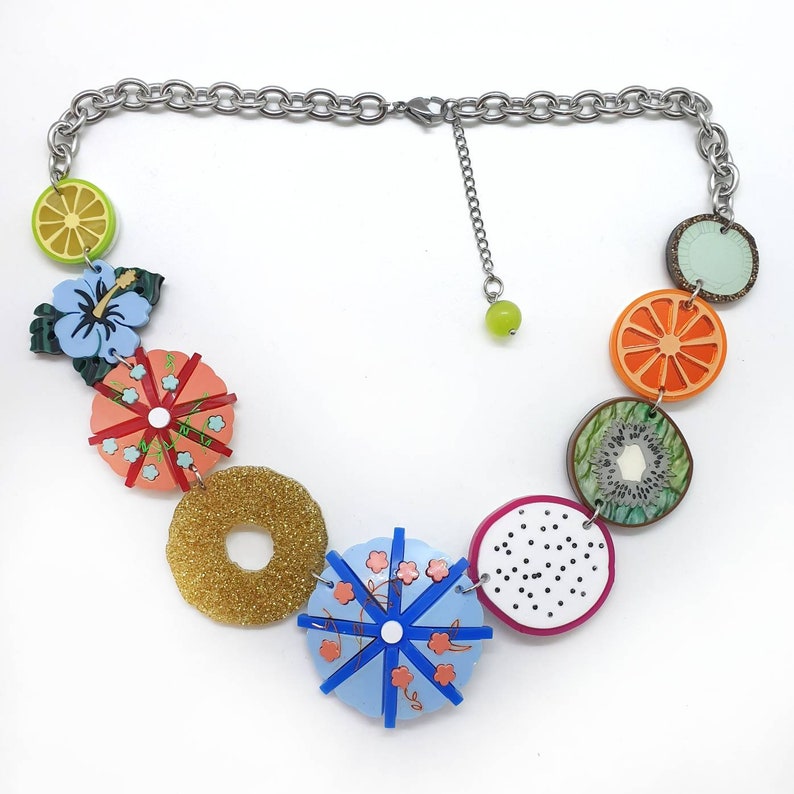 May include: A colorful fruit-themed necklace with a silver chain. The necklace features a variety of fruit slices, including orange, kiwi, and dragon fruit, as well as a pineapple and a hibiscus flower.