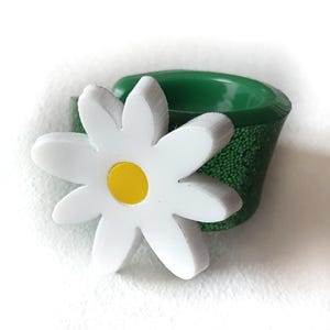 Può includere: Un fiore di margherita bianca con centro giallo è attaccato a un contenitore verde. Il contenitore ha un aspetto strutturato e maculato. Il fiore ha sei petali.