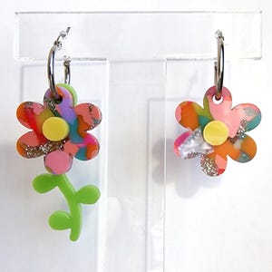 May include: A pair of silver hoop earrings with colorful flower charms. One charm is a single flower with a yellow center and the other is a flower with a stem and leaves.