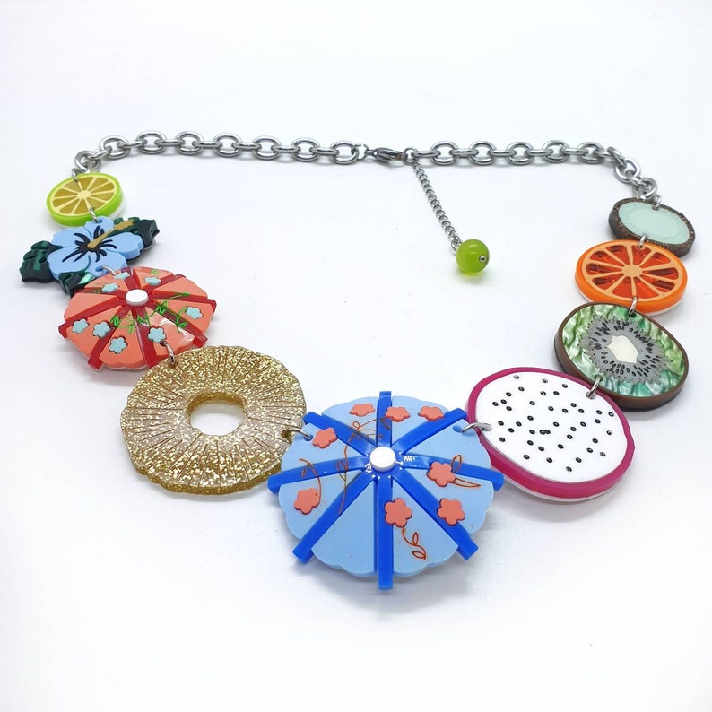 May include: A colorful necklace with a silver chain and various fruit and flower charms. The charms include a pineapple, a watermelon, a kiwi, a lime, a flower, and a donut.