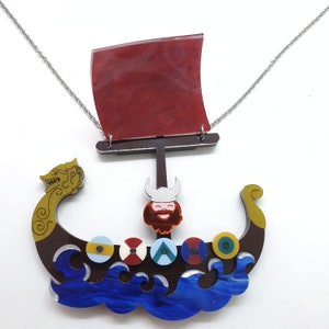 Può includere: Una collana a forma di nave vichinga rossa e marrone con una figura di prua barbuta e una vela rossa. La nave è circondata da onde blu e ha una testa di drago dorata a prua.