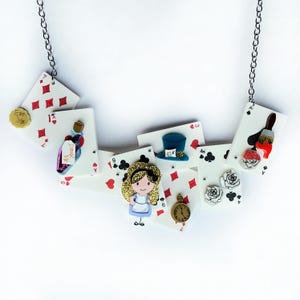 Op de afbeelding: Een grillige ketting met speelkaarthangers en bedels met het thema Alice in Wonderland. De bedels omvatten een fles met het opschrift "Drink Me", een hoed van de Mad Hatter, een zakhorloge en een Alice-figuur. De ketting is opgehangen aan een zilveren ketting.