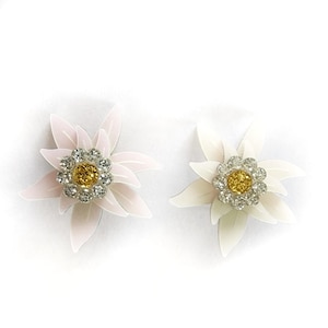 Può includere: Due spille a forma di fiore. Una è rosa chiaro, l'altra bianca. Ognuna ha un gruppo centrale di cristalli gialli e trasparenti, circondati da petali bianchi. Le spille sono su uno sfondo bianco.