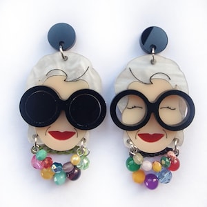Può includere: Un paio di orecchini pendenti con un viso di cartone animato con occhiali da sole neri e una frangia di perline colorate.