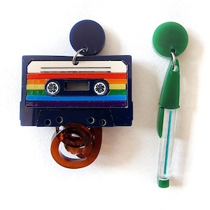 Può includere: Un paio di orecchini colorati. Un orecchino ha la forma di una cassetta con un design arcobaleno e una bobina di nastro marrone. L'altro orecchino è un tubo verde e blu con un cerchio verde in cima.
