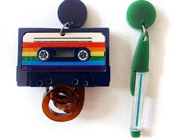 May include: A pair of colorful earrings. One earring is shaped like a cassette tape with a rainbow design and a brown tape spool. The other earring is a green and blue tube with a green circle at the top.