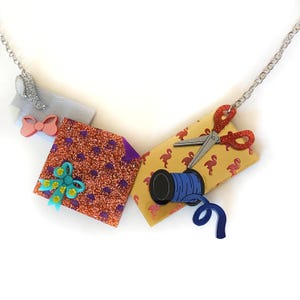 Può includere: Collana con catena argentata con ciondoli a forma di confezione regalo, forbici e rocchetto di filo. La confezione regalo è arancione con pois viola e un fiocco turchese. Le forbici sono rosse e la bobina è blu.