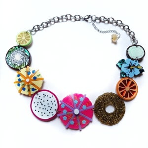 May include: A colorful necklace with fruit and flower charms. The necklace is made of silver chain and features a variety of charms, including a lime, kiwi, pineapple, dragon fruit, hibiscus flower, and orange slice.