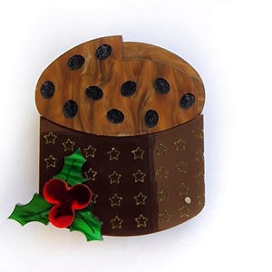 Può includere: Spilla a forma di torta di Natale marrone con una parte superiore marmorizzata e ritagli a forma di stella. La spilla presenta un rametto di agrifoglio con bacche rosse e foglie verdi, perfetta per le festività.