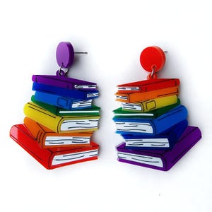 May include: A pair of rainbow-colored book stack earrings. Each earring features a stack of seven books in a rainbow color scheme, with the top book being purple and the bottom book being red. The earrings are made of acrylic and have a silver post.