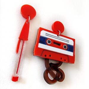 May include: A pair of red and white cassette tape earrings with a brown swirl design. The earrings are made of acrylic and have a vintage look.