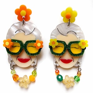 Può includere: Un paio di orecchini pendenti con un viso di cartone animato con occhiali da sole verdi, un sorriso rosso e un'acconciatura bianca. Gli orecchini sono decorati con perline e fiori colorati.