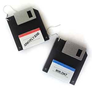 Puede incluir: Un par de pendientes de disquete negros con detalles metálicos plateados. Cada pendiente presenta una etiqueta blanca con texto, uno con "ARKYFLY.EXE" y el otro "80S.DAT". Los pendientes tienen cierres de gancho de plata.