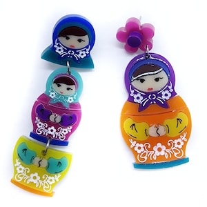 May include: Colorful Matryoshka doll earrings. One earring features a flower stud with a purple center. The other earring has three stacked dolls in blue, teal, pink, and yellow. Each doll has a painted face and floral accents.