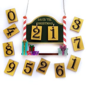 Puede incluir: Un letrero de cuenta regresiva navideña con una pizarra verde, postes a rayas rojas y blancas y una cadena para colgar. El letrero dice "DAYS TIL CHRISTMAS" y muestra "21". Incluye etiquetas numeradas y cajas de regalo decorativas.