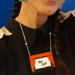 May include: A silver chain necklace with a red and white movie theater curtain pendant. The pendant has the words "The End" on it. The necklace is worn by a person with a black shirt and a braid. The text "Parure cinema" is written in purple below the pendant.