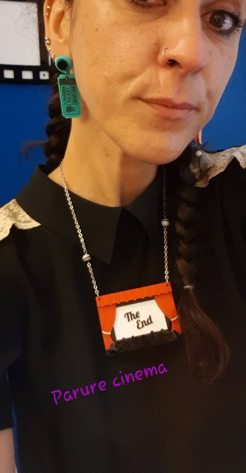 May include: A silver chain necklace with a red and white movie theater curtain pendant. The pendant has the words "The End" on it. The necklace is worn by a person with a black shirt and a braid. The text "Parure cinema" is written in purple below the pendant.