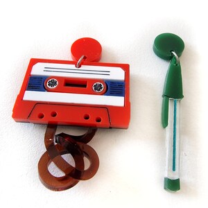 May include: A pair of colorful earrings. One earring is shaped like a cassette tape with a red body, white label, and blue accents. The other earring is a green and white pen-shaped design.