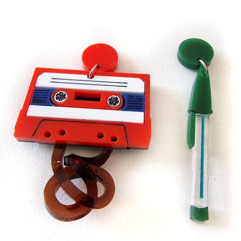 May include: A pair of colorful earrings. One earring is shaped like a cassette tape with a red body, white label, and blue accents. The other earring is a green and white pen-shaped design.