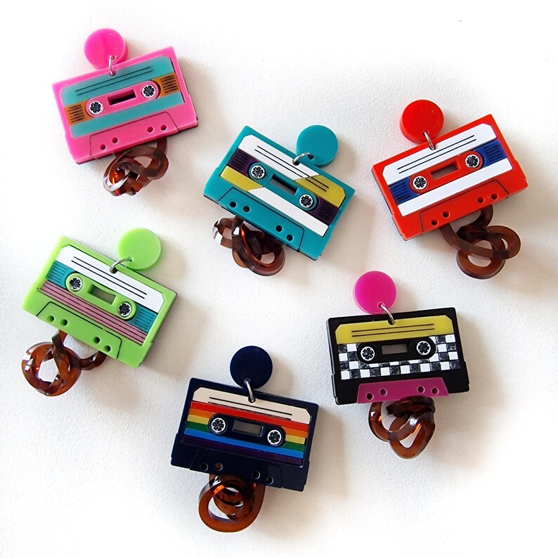 May include: Six pairs of colorful cassette tape earrings. Each pair features a different design and color combination, including rainbow, green, blue, red, and black and white.