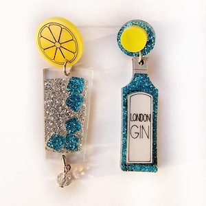 Op de afbeelding: Een paar oorbellen van blauwe en zilveren glitter. De ene oorbel is een glas gin met een citroenschijfje erop. De andere oorbel is een fles gin met de tekst "LONDON GIN" op het etiket.