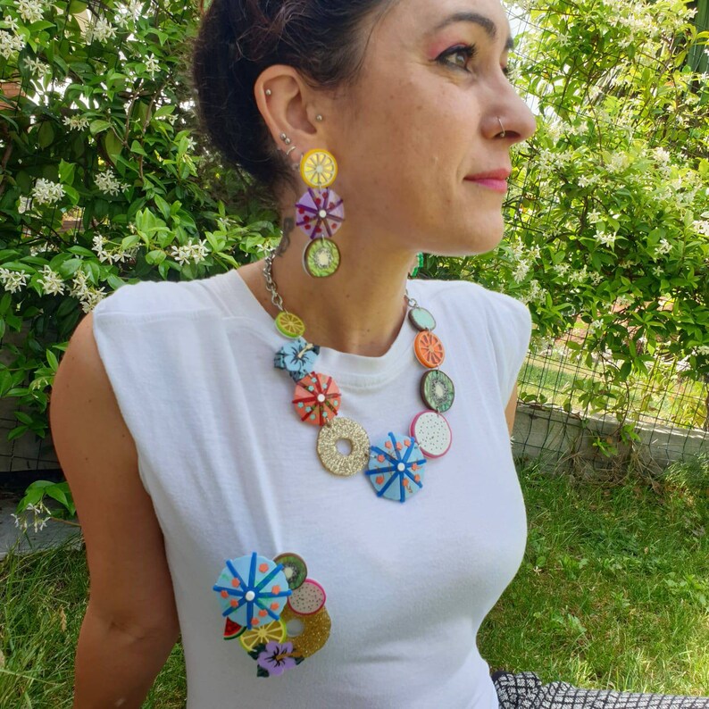 May include: A white t-shirt with a colorful fruit necklace and matching earrings. The necklace features a variety of fruits, including lemons, oranges, and kiwi. The earrings are a single fruit design.
