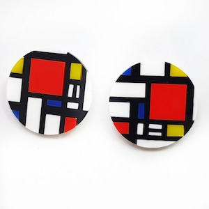 May include: Two round earrings with a Mondrian-inspired design. Each earring features a geometric pattern of red, yellow, blue, black, and white shapes. The earrings are set against a white background, highlighting the bold colors and abstract design.