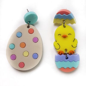 Può includere: Un paio di orecchini colorati a tema pasquale. Un orecchino presenta un uovo marrone chiaro con pois multicolori. L'altro orecchino presenta un pulcino giallo in piedi su un uovo blu, rosa e verde.