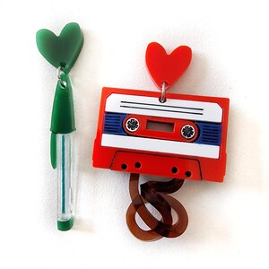 May include: A pair of earrings featuring a red cassette tape with a brown tape loop and a red heart charm, and a green pen with a green heart charm.