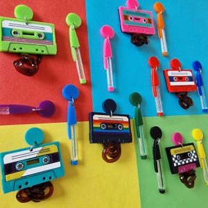 May include: Assortment of colorful cassette tape earrings with dangling charms. The earrings feature various colors and designs, including green, blue, pink, red, and black. Each earring has a cassette tape charm with a different design, such as a rainbow, checkered pattern, and a solid color.