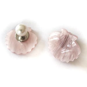 Può includere: Due spille a forma di conchiglia. Una è di colore rosa chiaro con una perla e uno spillo color argento. L'altra è a forma di conchiglia rosa chiaro. Entrambe sono su sfondo bianco.