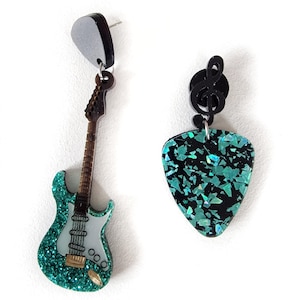 Puede incluir: Un par de pendientes colgantes con una guitarra eléctrica verde con purpurina y una púa de guitarra verde con purpurina con un dije de clave de sol negro.