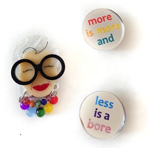 Könnte beinhalten: Eine Sammlung von drei Anstecknadeln. Eine Anstecknadel ist eine Brosche in Form eines Frauengesichts mit großer schwarzer Brille, rotem Lippenstift und einer bunten Perlenkette. Die anderen beiden Anstecknadeln sind rund mit dem Text "more is more and" und "less is a bore".